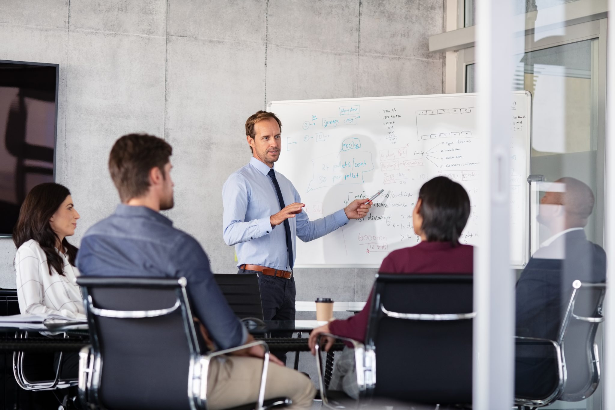 Mature businessman giving presentation to his colleagues in modern office. Formal leader presenting new project to business partners in conference room using white board. Businessman in a meeting showing business progressions in boardroom.